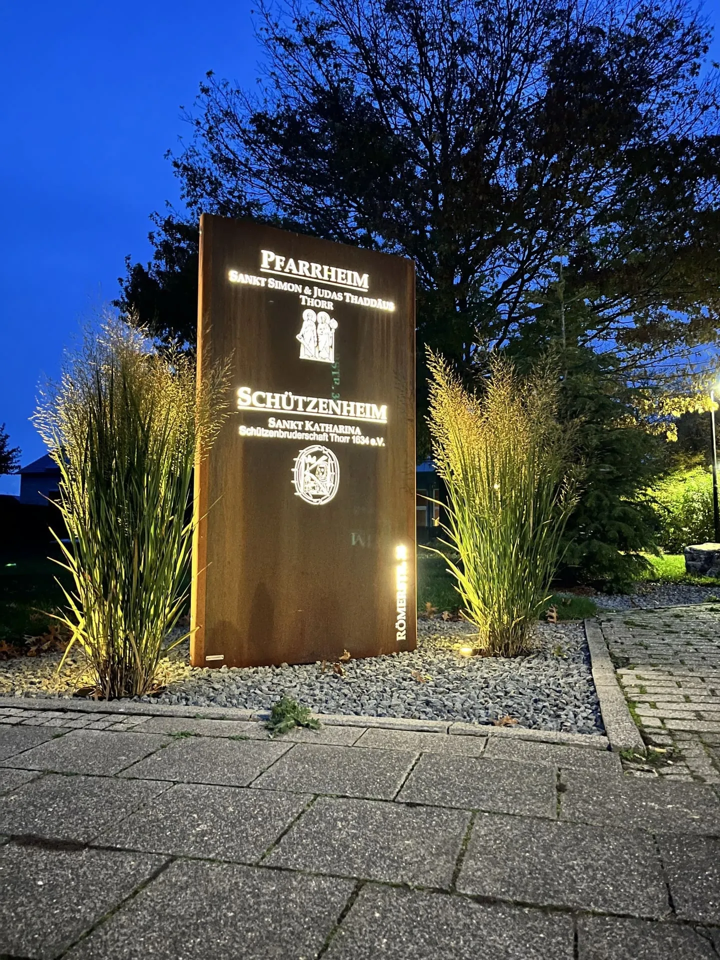 Beleuchtete Stele „Pfarrheim / Schützenheim" bei Dämmerung, eingerahmt von Gräsern
