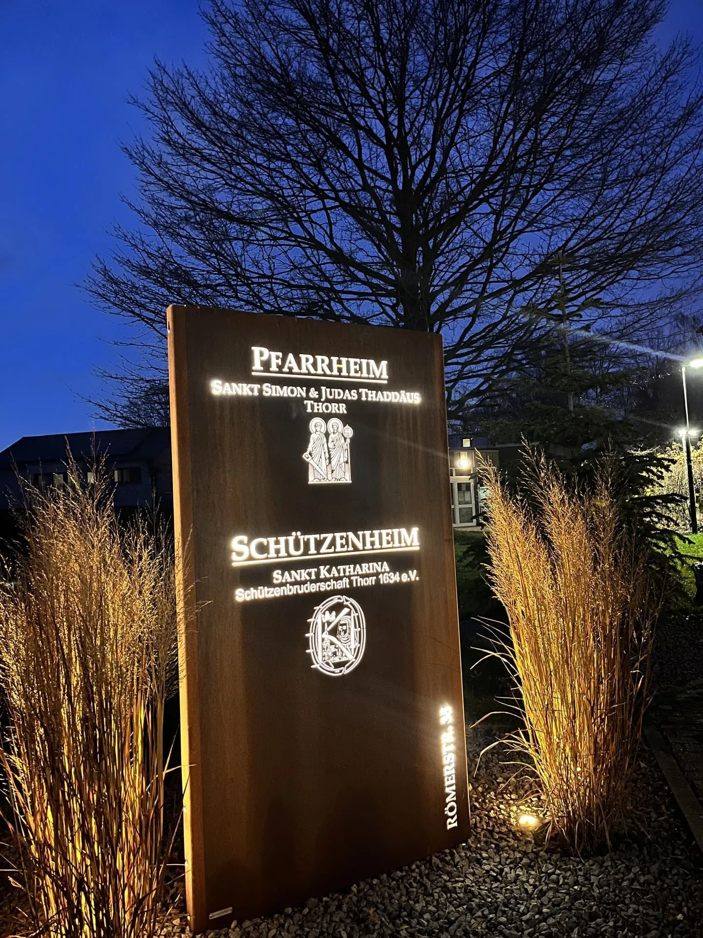 Beleuchtetes Hinweisschild mit Wappen von Pfarrgemeinde und Bruderschaft bei Nacht