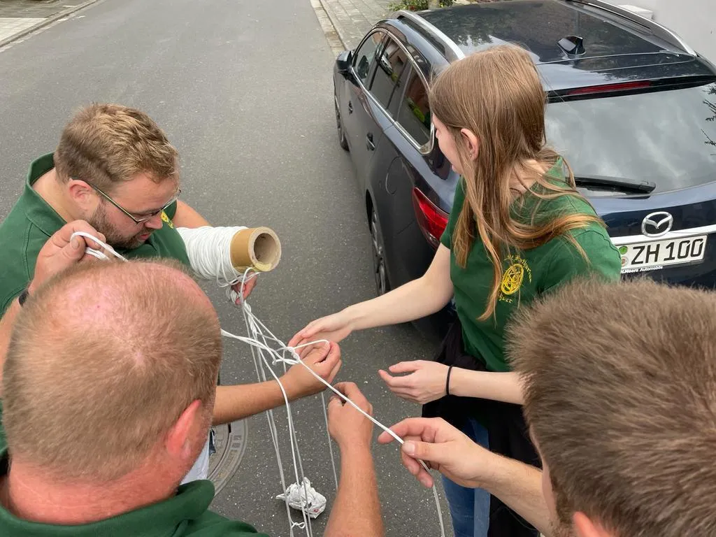 Schützenbrüder und -schwestern fädeln gemeinsam Schnüre zum Wimpelaufhängen, im Hintergrund ein dunkles Auto