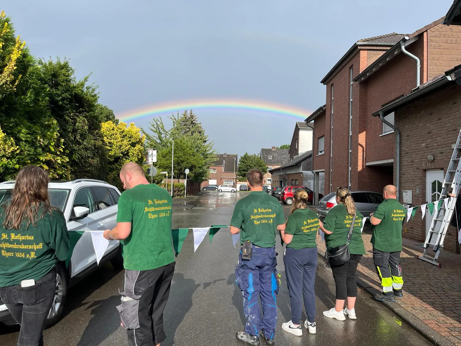 Helfer mit Wimpelketten auf nasser Dorfstraße, ein Regenbogen über den Häusern nach dem Schauer