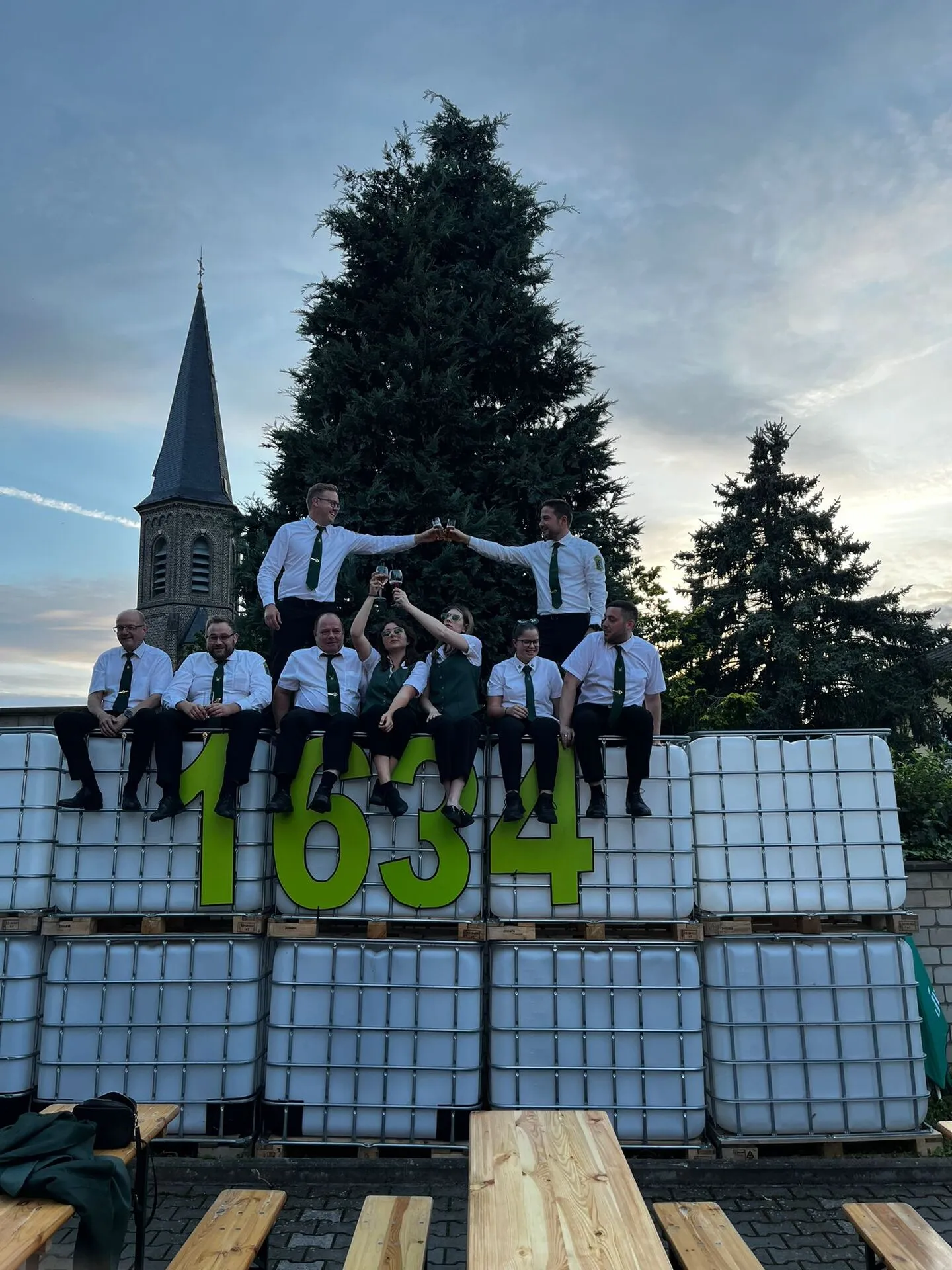 Junggesellen-Zug auf der „1634"-Bühne aus IBC-Containern, zwei stehen oben und stoßen mit Flaschen an, Kirchturm im Abendrot