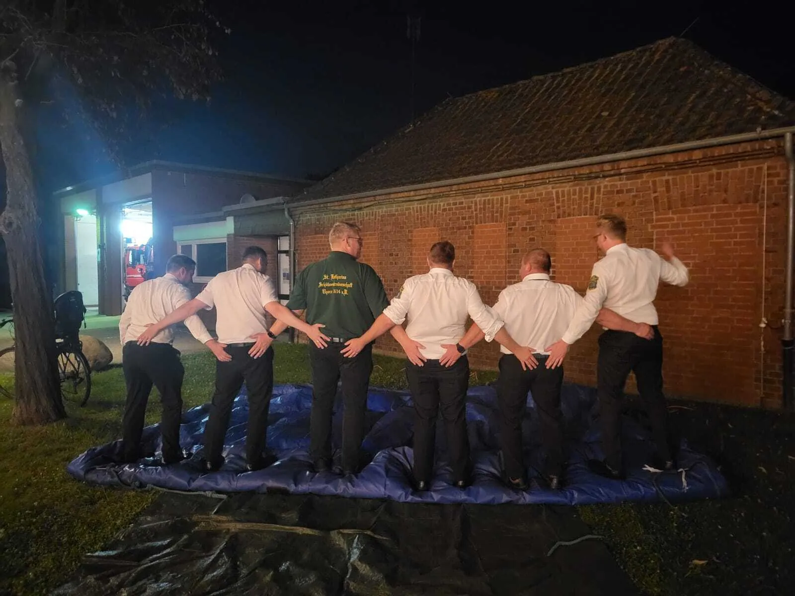 Sechs Schützenbrüder stehen abends von hinten Schulter an Schulter auf einer blauen Plane, im Hintergrund ein Backsteingebäude