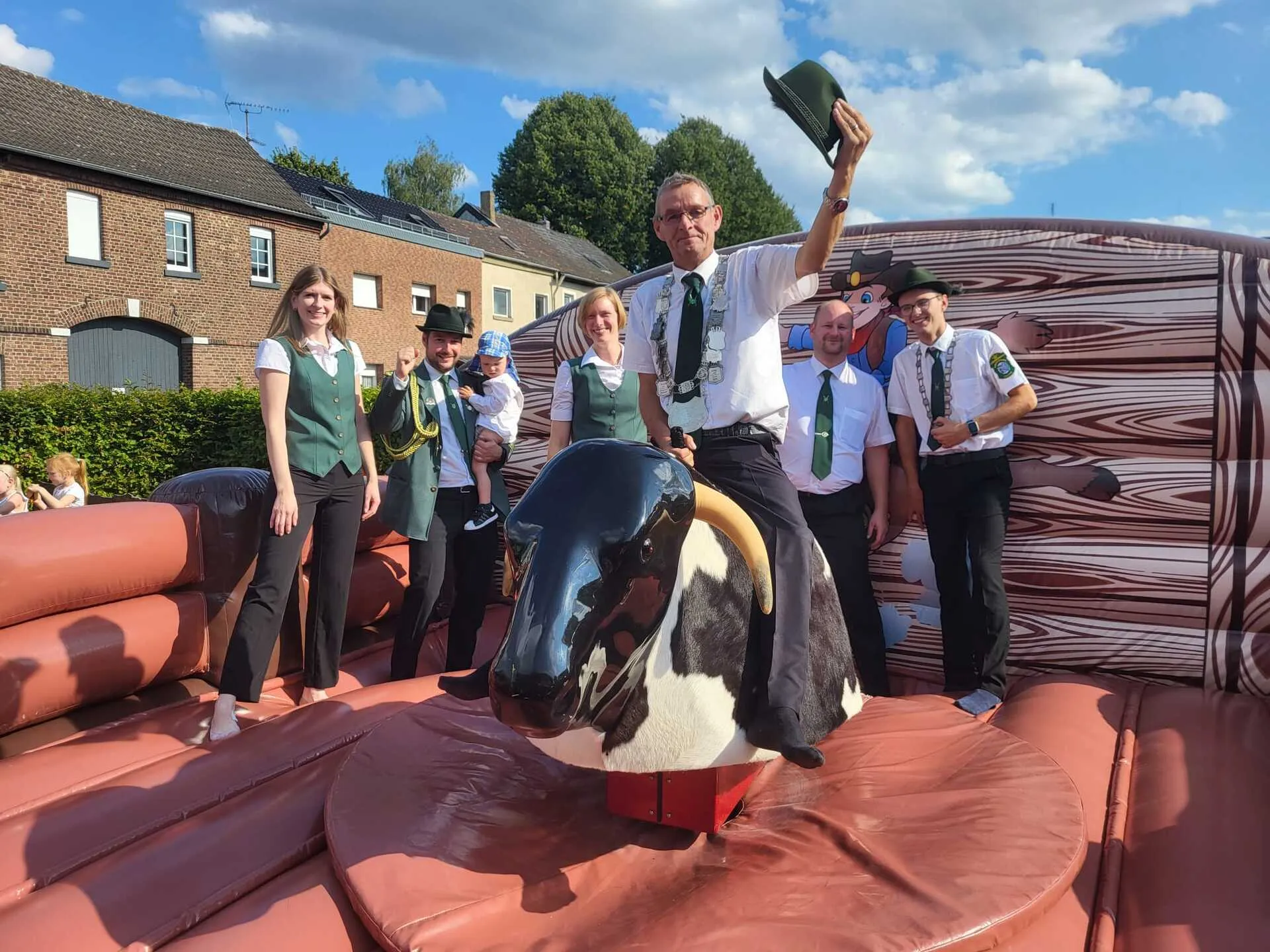 Schützenkönig lacht auf dem Bullriding-Bullen, Hut in der Hand, Bruderschaft im Hintergrund