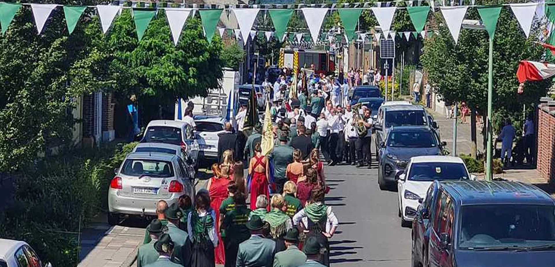Festumzug durch eine geschmückte Straße mit Menschen in Trachten und Musikgruppe, begleitet von Zuschauern und parkenden Autos